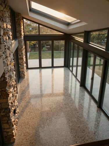 Overhead perspective of an empty dining volume in a modernist villa, terrazzo floor reflecting a skylight, dark steel window frames, stone wall detail at left edge, golden hour light entering at a low angle through clerestory windows