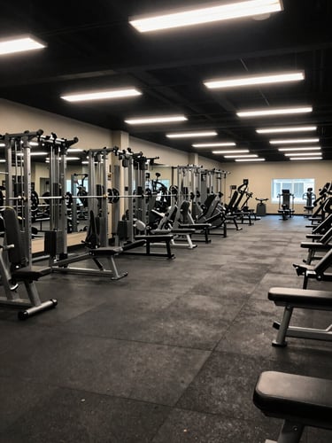 Wide environmental shot of a completed commercial gym interior, overhead fluorescent lighting, full rack configuration visible left to right, rubber flooring with equipment spacing that shows a deliberate floor plan, no people present