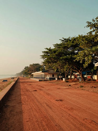 Wide environmental landscape shot of Prantik station area with red soil paths and lush green trees under a soft coastal haze, 35mm lens