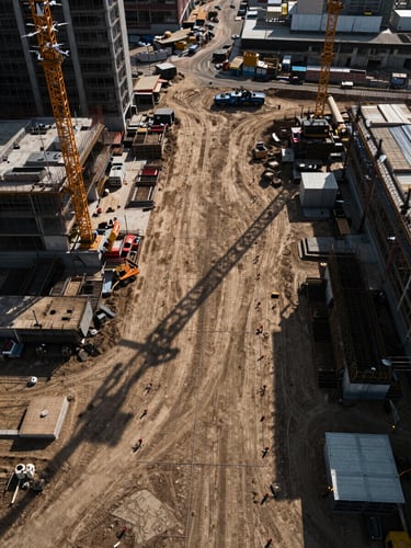 Wide panoramic view from above a major urban construction site, survey drone visible in the upper left corner, grid of survey markers and reference flags across raw compacted earth, long shadows of cranes and equipment in late afternoon light — rich texture of disturbed soil and concrete formwork