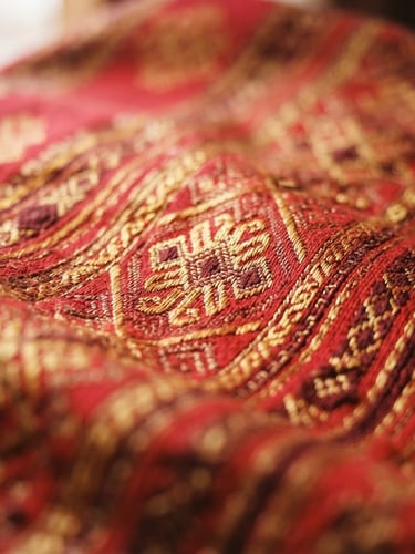 Close-up of intricate hand-woven Bhutanese textile with rich crimson and gold threads, warm natural window light, shallow depth of field, 35mm