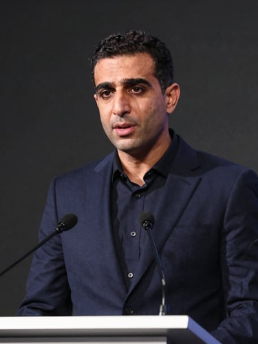 Medium-close environmental portrait of Karim speaking at a high-level conference podium, crisp natural lighting, deep navy tones, 35mm
