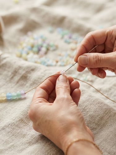 Macro close-up of hands mid-assembly, threading soft pastel glass beads onto a cord over a textured linen cloth, warm diffused daylight