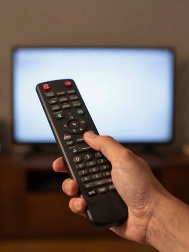 Close-up of a hand pointing a TV remote directly at a glowing television screen in a dim living room, tungsten warm light from the screen illuminating the hand, framed tight on the device mid-press, left-side composition with dark ambient background