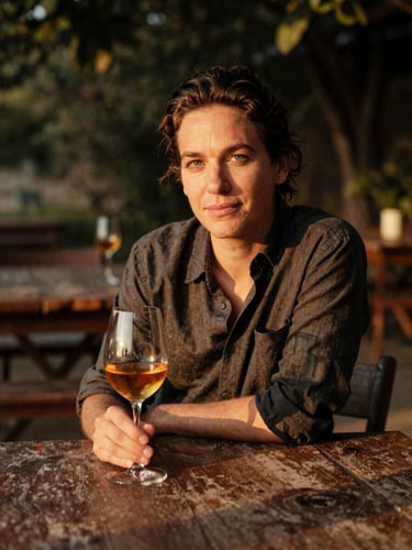 Environmental portrait of a culinary writer sitting at a rustic outdoor table, holding a glass of amber wine, warm golden-hour light