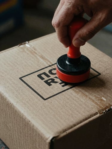 Close-up of a hand stamping a cardboard shipping box with a bold black ink brand seal, gritty workshop background.