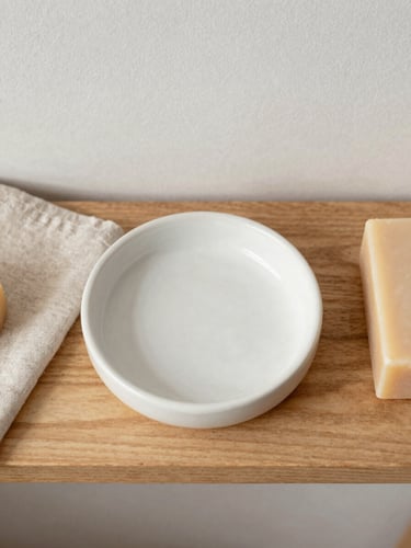 Overhead flat-lay of a wooden shelf with a ceramic soap dish, textured linen cloth, and a bar of body soap under soft morning light.