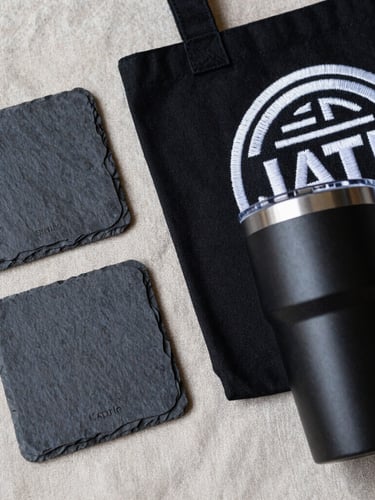 Macro flat-lay of finished custom products: a laser-engraved slate coaster, a heavy black canvas tote with dense white embroidery, and an anodized matte black tumbler, all arranged on a raw linen background
