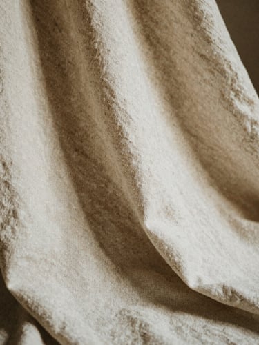 Extreme close-up of heavy organic linen fabric drape, soft natural afternoon light, deep shadows, warm bone and espresso tones, editorial 35mm photography