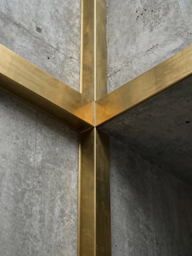 Macro abstract photograph of heavy architectural concrete meeting polished brass, sharp geometric lines, dramatic low-angle lighting with deep shadows and golden highlights.