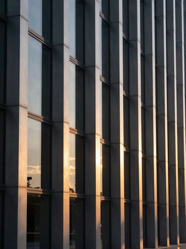 A dramatic, high-contrast architectural photograph of modern steel and glass columns reflecting a deep golden-hour sky. Sharp highlights, deep shadows, clean geometric lines.