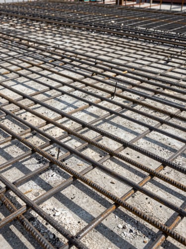 Close-up ground-level view of dense rebar grid on a concrete slab at an active construction site, natural north-facing daylight, geometric shadow patterns across the steel, no people, material texture dominant