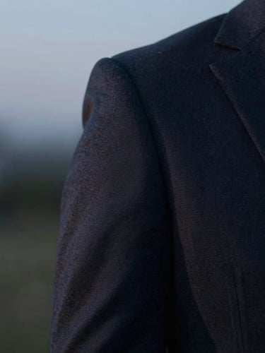A close-up of a sharp, tailored dark suit sleeve with a subtle earpiece visible in the soft-focus background, cool twilight tones