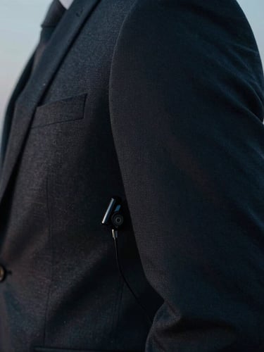 Cinematic close-up of a sharp, tailored dark suit sleeve with a subtle, low-profile earpiece visible, cool twilight tones, high contrast.