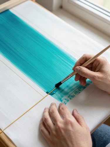 Overhead shot of hands holding a fine paintbrush, guiding vibrant teal dye along a golden resist line on stretched white silk, bright natural window light, 35mm