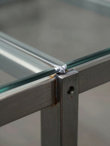 Extreme close-up of a steel-to-glass table joint under raking sidelight, weld seam and edge chamfer in sharp focus, cool neutral studio light, no props, surface texture of brushed steel visible