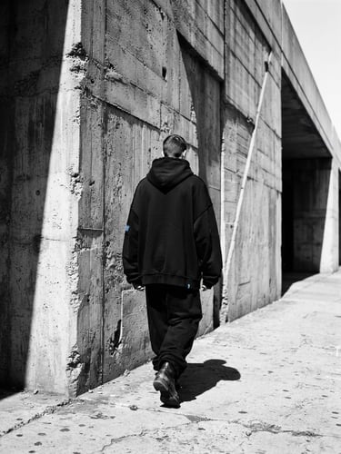 Low-angle street photography of a concrete brutalist wall, sharp harsh midday sun shadows, a model from behind wearing a black heavyweight hoodie with raw edges, 35mm film style