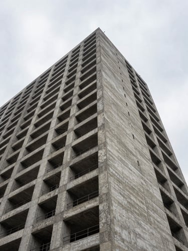 Low-angle heroic shot of a completed mid-rise building against an overcast sky, emphasizing raw concrete and rigid geometric shadows.