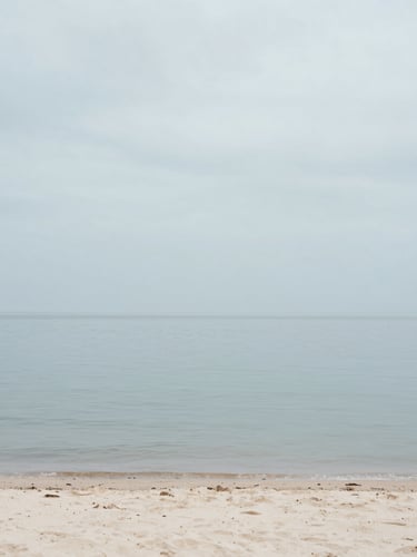 Tranquil coastal shoreline with calm water, soft overcast gray sky, pale sand, minimalist editorial photography, 35mm lens