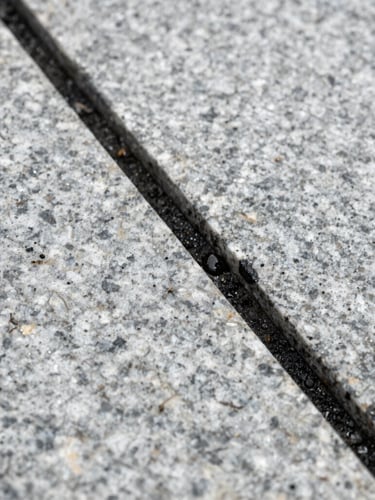 Macro straight-on shot of weathered exterior stone tiles, a sharp diagonal line separating deep black grime from pure light gray granite, water droplets glistening under bright overcast sky.