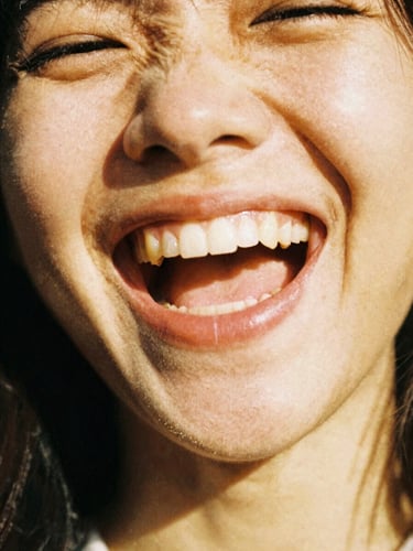 Close-up of a person laughing in harsh direct sunlight, strong shadows, saturated golden tones, heavy grain, analog 35mm style