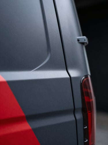 Extreme close-up macro of a precision wrap edge on a dark graphite delivery van, matte vinyl surface transition meeting a crimson graphic stripe, cool studio lighting revealing zero-gap seam quality, black background