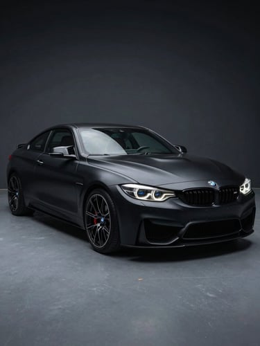 Wide studio shot of a matte black BMW M4 three-quarter front angle, full wrap with precision-cut edges, dark charcoal background, cool studio floor lighting revealing vinyl surface texture and depth, no glare, shallow depth of field on front quarter panel