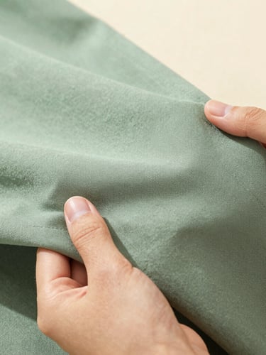 Close-up of a high-quality sage green fabric texture being held gently by female hands, showing neat stitching and premium weave, soft natural lighting, shallow depth of field.