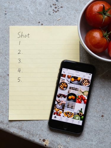 A flat lay on a worn kitchen prep table: a shot list on yellow notepad paper, a phone showing a draft Instagram grid, a small bowl of heirloom tomatoes, all under cool north-facing daylight, no artificial light