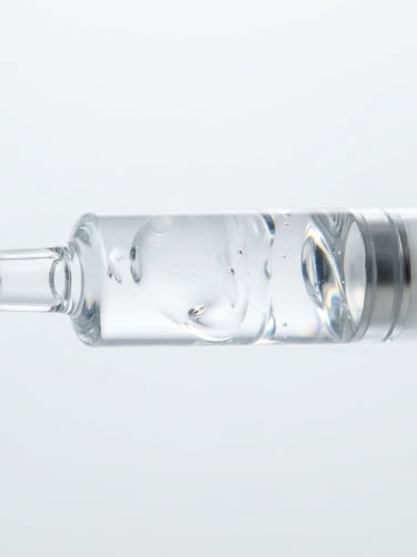 Macro close-up of a high-purity clear gel syringe showing viscosity, bright high-key studio light with sharp refractions through glass, clinical white and silver tones.