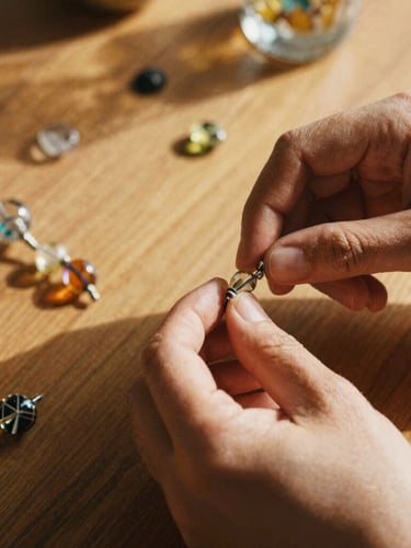 Close-up of hands working with intricate glass beads on a wooden table, warm editorial photography, natural golden-hour daylight, rich earthy tones, shallow depth of field, 35mm film texture.