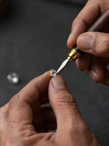 Extreme close-up portrait of a master artisan's hands using traditional tools to delicately press pure gold foil around an uncut Polki diamond, warm studio lighting with a deep chiaroscuro effect, dark charcoal background.