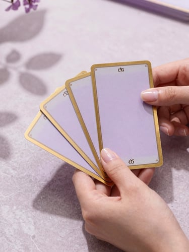 Hands with elegant clean manicure gently holding gold-rimmed tarot cards over a stone table, soft lilac morning light, botanical leaf shadows, 35mm