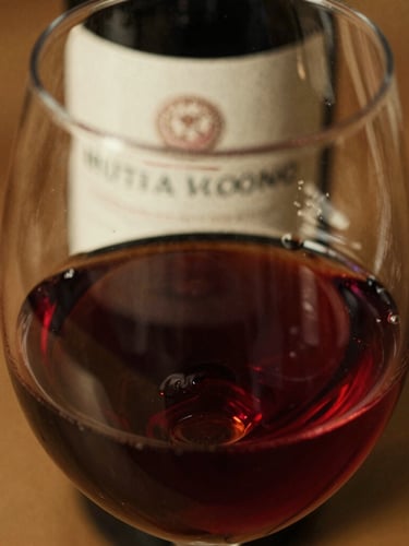 Macro shot of red wine swirling in a clear glass, warm golden hour light, rich burgundy tones, textured paper label of a wine bottle slightly visible in the background, 35mm film look.