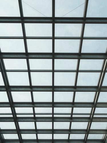 Structured architectural perspective looking straight up at a glass ceiling with clean geometric steel frames, cool daylight.