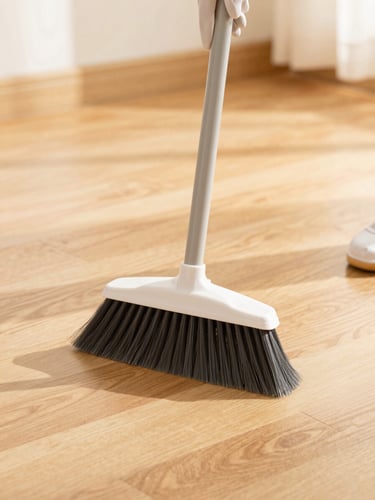Close-up of hands in protective gloves holding a clean broom, sweeping dust off a warm wooden floor in a bright sunlit room, sharp focus, editorial style.