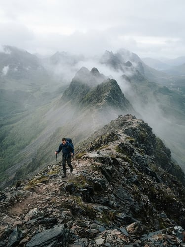 A hiker navigating a narrow mountain ridge under misty morning light, desaturated forest greens and slate grays, wide-angle lens