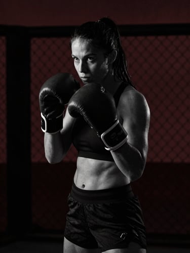 Black and white portrait of a female fighter in a dark gym, harsh side light, chain-link background, dramatic low-key lighting, 35mm lens