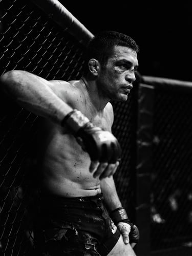 Dramatic high-contrast black and white portrait of a focused MMA fighter leaning against a chain-link cage, harsh directional key light, deep shadows, 35mm lens
