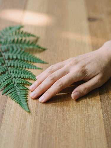 A close-up of a person's hand gently resting on a textured wooden table next to a small green fern, soft morning light, warm organic tones, 35mm editorial photography