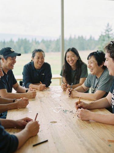 Candid 35mm film photo of diverse community members laughing around a large wooden studio table, warm overcast daylight, paint-stained hands holding charcoal pencils.