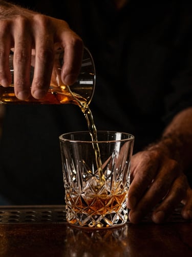 Dark moody macro shot of a bartender's hands in calm motion pouring a golden amber liquid from a mixing glass, shallow depth of field, low-key lighting