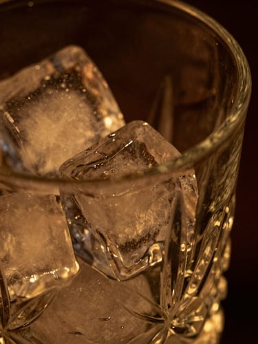 Close-up macro of a crystal mixing glass, clear ice block reflecting low-key warm amber light, dark moody background, shallow depth of field, 35mm