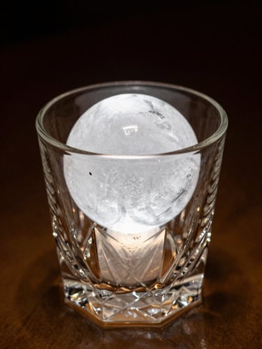 Macro shot of a perfect hand-carved clear ice sphere sitting in a heavy crystal rocks glass, dark warm amber lighting, dramatic shadows, 35mm