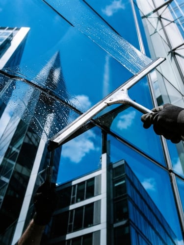 Close-up of a professional squeegee pulling water off a large clean glass window, bright daylight, electric blue sky reflections, sharp architectural lines, 35mm.