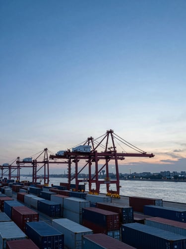 Wide panoramic view of a commercial container port at dawn, cranes silhouetted against a cool steel-blue sky, rows of shipping containers receding to the horizon on the left, open water visible to the right, crisp shadow-rich industrial light