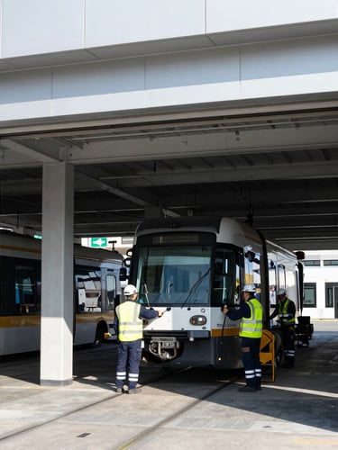 High-contrast daylight shot of a modern transit maintenance depot, clean architectural framing, technicians in safety gear inspecting a tram undercarriage, industrial cool tones