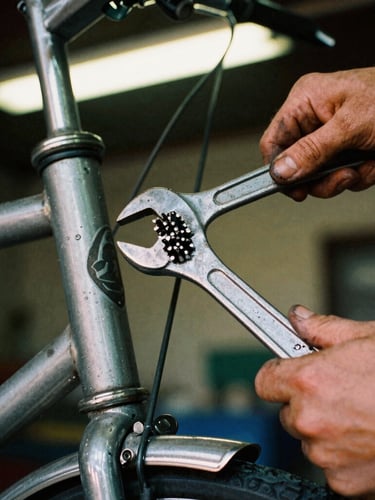 Close-up of grease-stained hands holding a heavy wrench over a vintage bicycle frame, warm raw workshop light, high contrast, 35mm film grain
