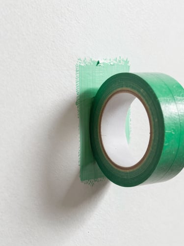 Macro shot of a painter peeling back crisp green painter tape, revealing an immaculate, razor-sharp paint line on a gallery-white wall, bright natural daylight