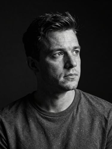 High-contrast black-and-white portrait of Russell James in low-key chiaroscuro lighting, looking thoughtful against a dark background.